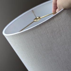 May include: A cylindrical lamp shade featuring a light gray, textured fabric exterior. The interior structure consists of a gold-toned metal spider fitter with three spokes meeting at a central ring, designed for attachment to a lamp base.