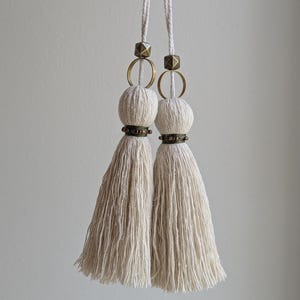 May include: Two off-white cotton tassels suspended from matching white cords. Each tassel features a round wooden bead, a circular brass ring, and a faceted brass cube bead at the top, with a band of small dark metal beads encircling the center.