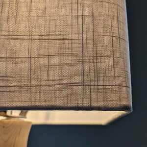 May include: Close-up of a beige lampshade with a grid pattern. The lampshade is rectangular and made of a textured fabric. The base of the lamp is light-colored wood, and the lamp is turned on, emitting a soft glow.