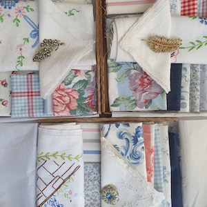 May include: A collection of vintage fabric scraps in various patterns and colours, including floral, plaid, and striped designs. The fabrics are arranged in a wooden box, with some of the pieces folded and others laid flat. The fabrics are mostly white, blue, and red.