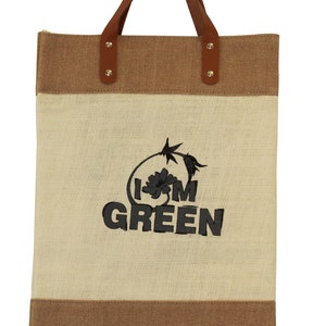 Puede incluir: Una bolsa de mano marr&oacute;n y beige con asas de cuero. La bolsa tiene un dise&ntilde;o bordado que dice "I'm Green" con una peque&ntilde;a flor.