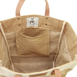 Puede incluir: Una bolsa de tote de yute marr&oacute;n con un forro blanco y un peque&ntilde;o bolsillo interior. La bolsa tiene asas de cuero y una etiqueta con el texto "Made in India" y un gr&aacute;fico de globo terr&aacute;queo.