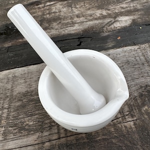 May include: A white ceramic mortar and pestle set. The mortar is a round bowl with a pouring spout, and the pestle is a cylindrical shape with a rounded end. The set is on a weathered wooden surface.