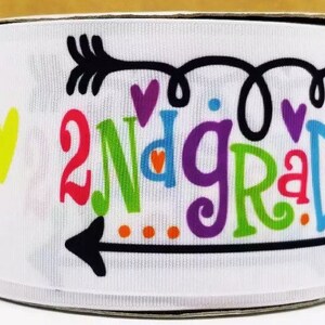 以下が含まれることがあります： 矢印とハートが描かれたカラフルなデザインの白いリボンで、「2nd Grade」と書かれています。