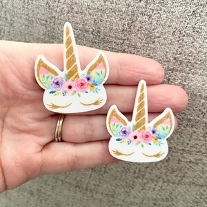 May include: Two white unicorn head shapes with gold horns and floral crowns. The flowers are pink, purple, and yellow. The unicorn eyes are closed.