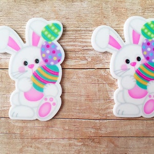 May include: Two white bunny rabbit figurines holding colorful Easter eggs. The bunnies are holding the eggs in their front paws. The eggs are decorated with stripes, polka dots, and swirls.