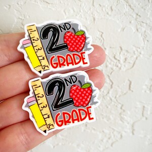 May include: Two gray and white stickers with a red apple and a yellow pencil with a ruler. The stickers say "2nd Grade" in black text.