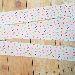 May include: White ribbon with a repeating pattern of colourful sprinkles. The ribbon is folded in thirds, showing the pattern from different angles.
