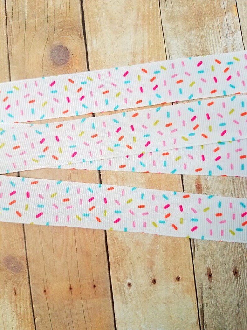 May include: White ribbon with a repeating pattern of colourful sprinkles. The ribbon is folded in thirds, showing the pattern from different angles.