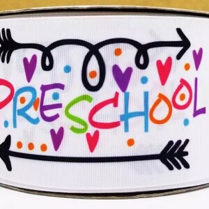 以下が含まれることがあります： 黒矢印とカラフルな文字で「PRESCHOOL」と書かれた白いリボン。ハートとドットが散りばめられています。
