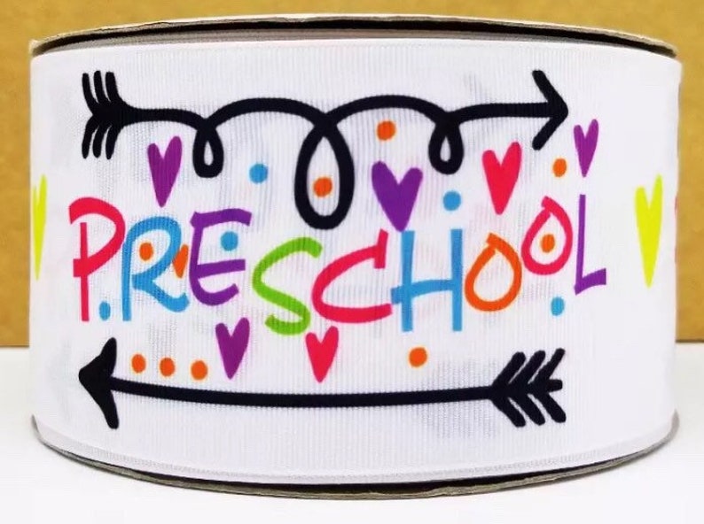 以下が含まれることがあります： 黒矢印とカラフルな文字で「PRESCHOOL」と書かれた白いリボン。ハートとドットが散りばめられています。