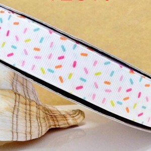 May include: White ribbon with a colourful sprinkle pattern. The ribbon is rolled up and sitting on a white seashell.