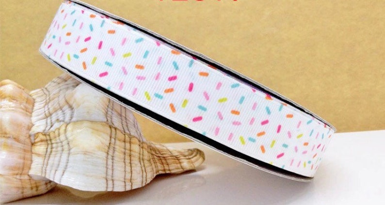 May include: White ribbon with a colourful sprinkle pattern. The ribbon is rolled up and sitting on a white seashell.