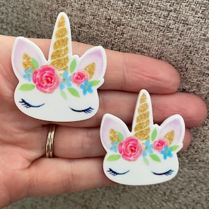 May include: Two white unicorn head shaped resin embellishments with gold glitter horns, pink and blue flowers, and closed eyes.