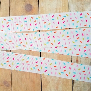 May include: White ribbon with a colourful sprinkle pattern. The ribbon is folded in thirds and laid out on a wooden surface.