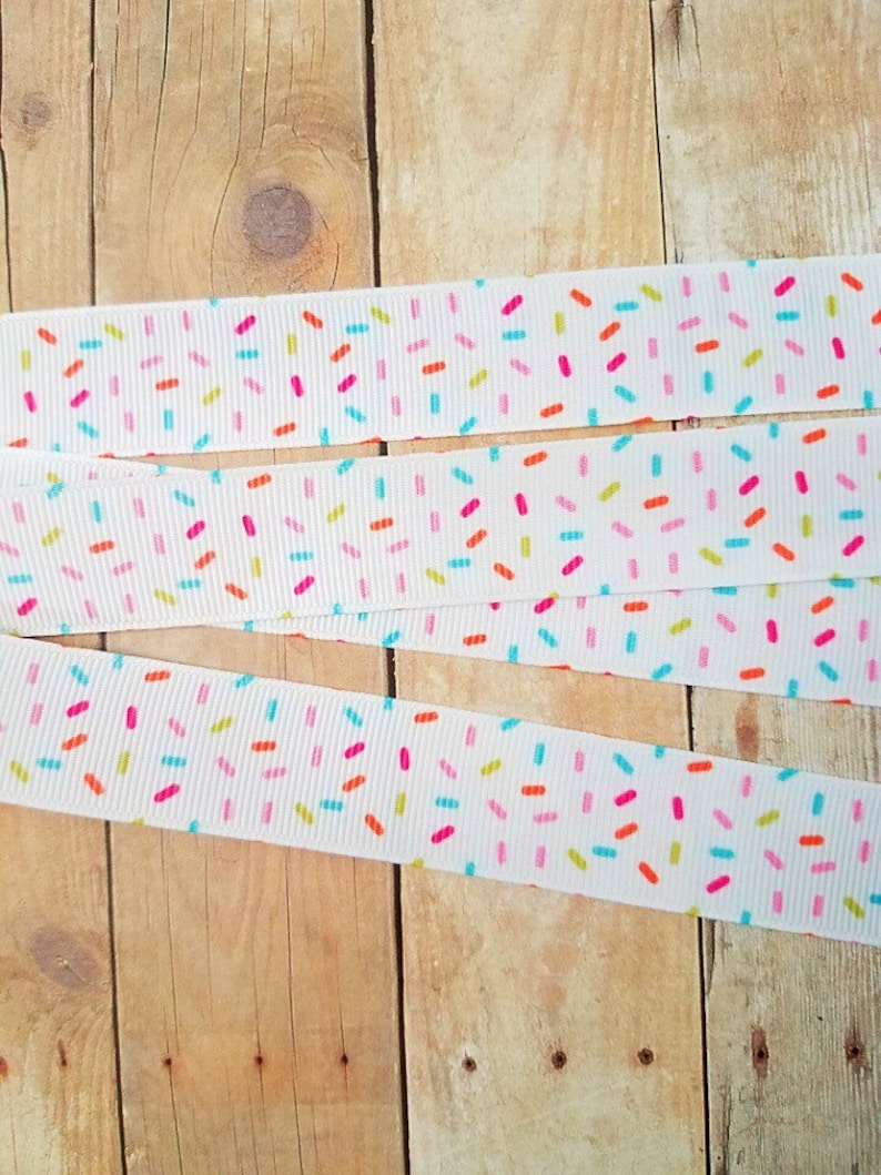 May include: White ribbon with a colourful sprinkle pattern. The ribbon is folded in thirds and laid out on a wooden surface.