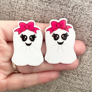 May include: Two white ghost-shaped ornaments with pink bows and black eyes and mouths. The ghosts have eyelashes and a smiling expression.