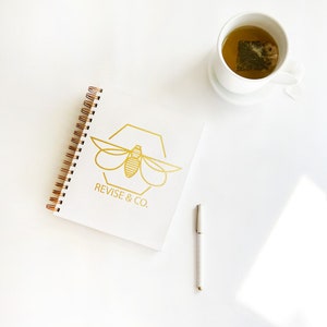 May include: A white spiral bound notebook with a gold bee logo and the text "REVISE & CO."  A gold pen and a white cup of tea are on a white surface.