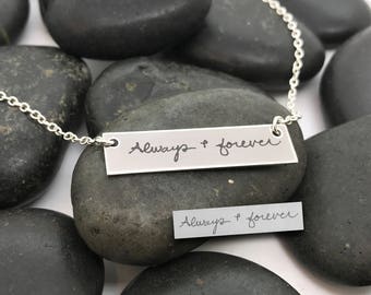 May include: A silver necklace with a rectangular pendant that says "Always + forever" in handwritten script. The necklace is on a bed of gray rocks.