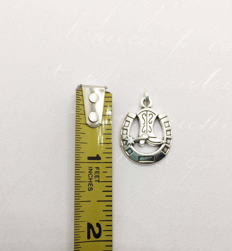 May include: Silver charm with a horseshoe design and a cowboy boot inside. The horseshoe has a star and a small circle on each side.