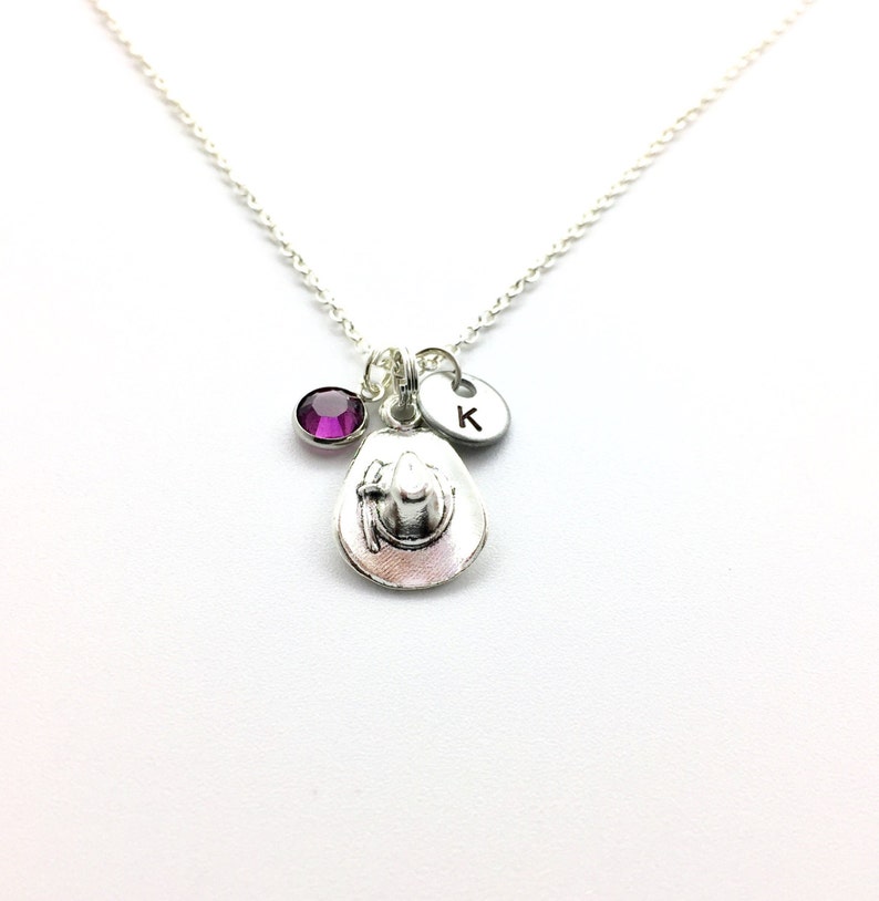 May include: A silver necklace with a charm of a cowboy hat, a purple gemstone, and a silver disc with the letter 'K' engraved on it.