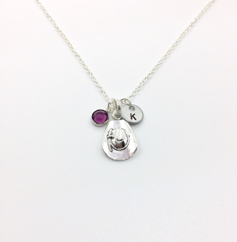 May include: A silver necklace with a charm of a cowboy hat, a purple gemstone, and a silver charm with the letter K.