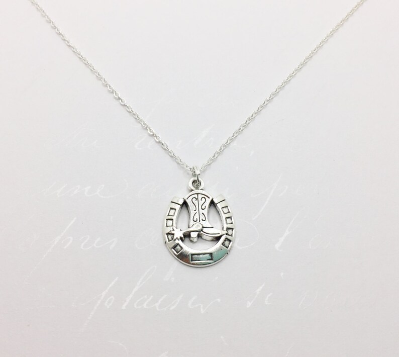 May include: A silver necklace with a horseshoe pendant featuring a cowboy boot and a star. The horseshoe is detailed with a textured design.