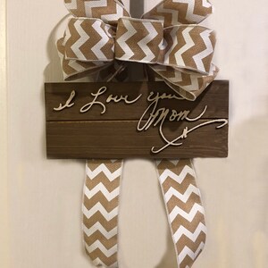 May include: A wooden sign with the text "I Love you Mom" written in cursive. The sign is decorated with a brown and white chevron patterned bow.
