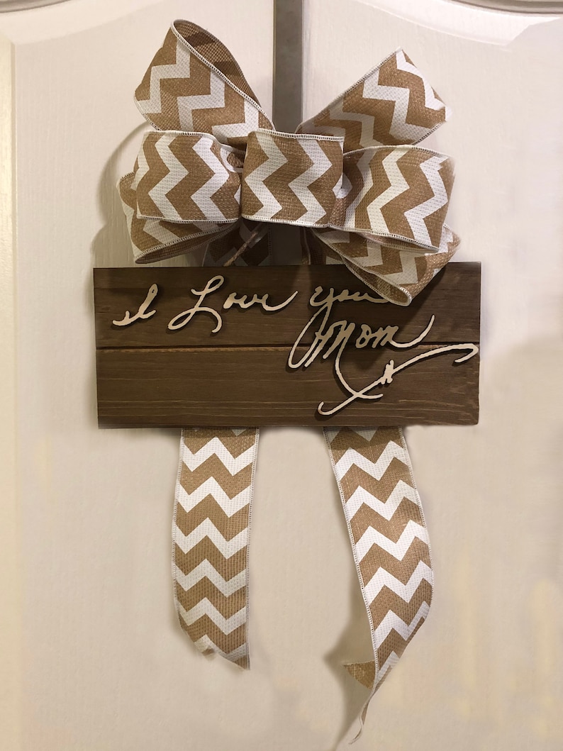 May include: A wooden sign with the text "I Love you Mom" written in cursive. The sign is decorated with a brown and white chevron patterned bow.