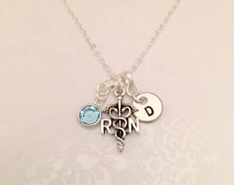 May include: A silver necklace with a charm featuring the medical symbol, the letters "RN", and a light blue crystal. The necklace also has a silver charm with the letter "D".