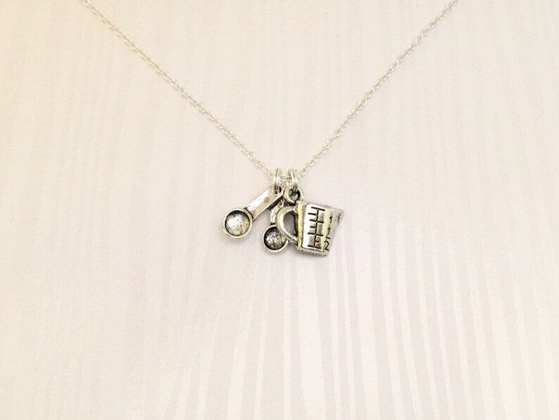 Puede incluir: Un collar de plata con un dije de una taza medidora y dos cucharas. El dije est&aacute; unido a una cadena de plata.