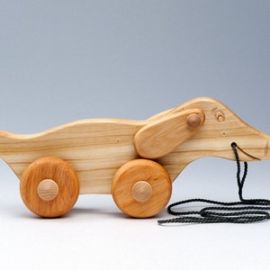 Può includere: Un giocattolo di legno a forma di bassotto con una corda per tirarlo. Il giocattolo ha una finitura marrone chiaro ed è su quattro ruote. Il bassotto ha un viso scolpito con un sorriso.