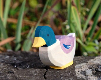 May include: A wooden duck toy with a blue head, yellow beak, purple body, and blue wing detail. The duck is standing on a gray, textured surface.