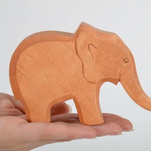 May include: A hand-carved wooden elephant figurine in a light brown color. The elephant is standing on all four legs with its trunk raised. The figurine is made of wood and has a smooth finish.