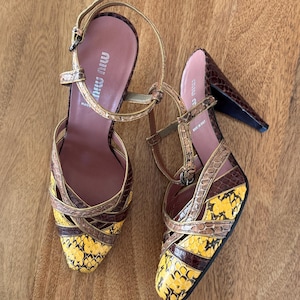 Może przedstawiać: Para brązowych i żółtych sandałów na obcasie ze skóry węża z złotymi akcentami. Sandały mają pasek wokół kostki i zapięcie na klamrę. Nazwa marki "Miu Miu" jest widoczna na wkładce. Wyprodukowano we Włoszech.