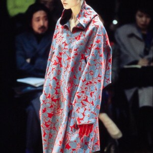 May include: A model walks the runway wearing a long, blue and red floral print coat with a high collar. The coat has a loose fit and is worn with purple tights.