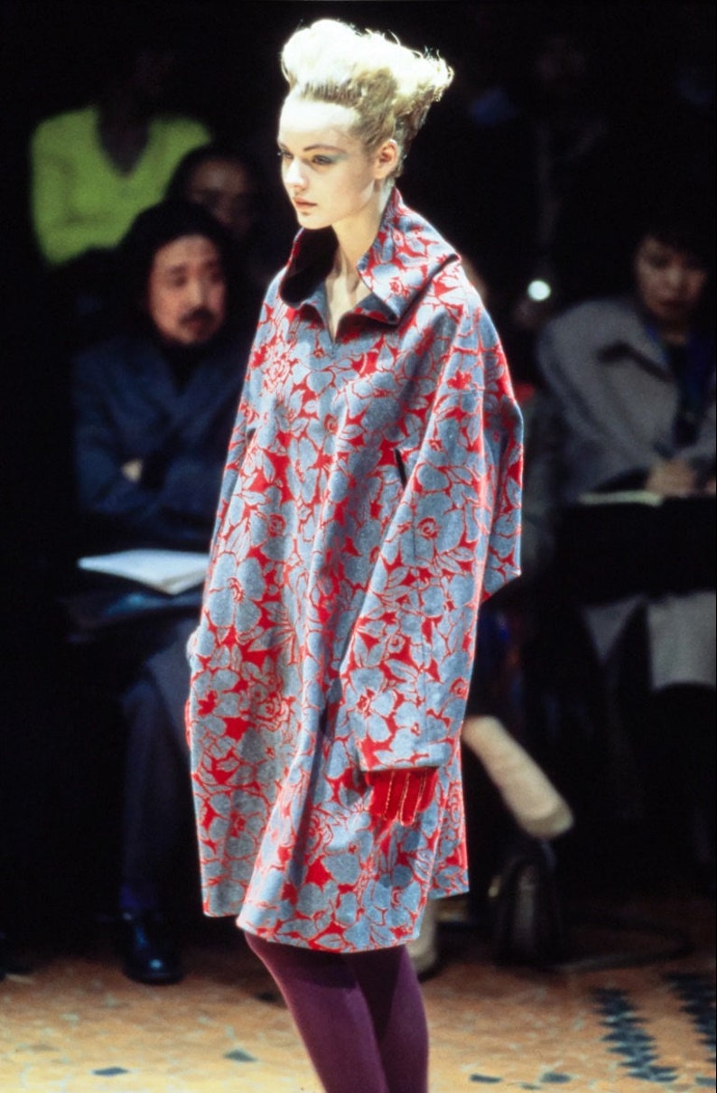 May include: A model walks the runway wearing a long, blue and red floral print coat with a high collar. The coat has a loose fit and is worn with purple tights.