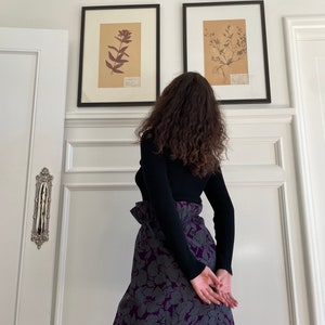 May include: A woman wearing a black mock neck sweater and a purple and gray floral skirt. The skirt has a high waist and a ruffled detail. The woman is standing in front of a white wall with two framed botanical prints.