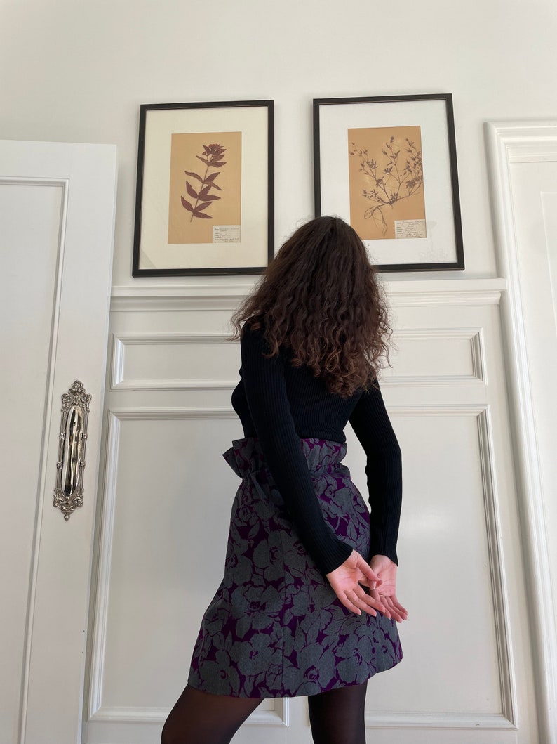May include: A woman wearing a black mock neck sweater and a purple and gray floral skirt. The skirt has a high waist and a ruffled detail. The woman is standing in front of a white wall with two framed botanical prints.