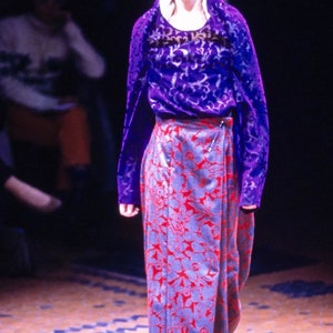 May include: A model wearing a purple and silver patterned top with a long, red and silver floral skirt. The skirt has a wrap-around style and is cinched at the waist with a belt.