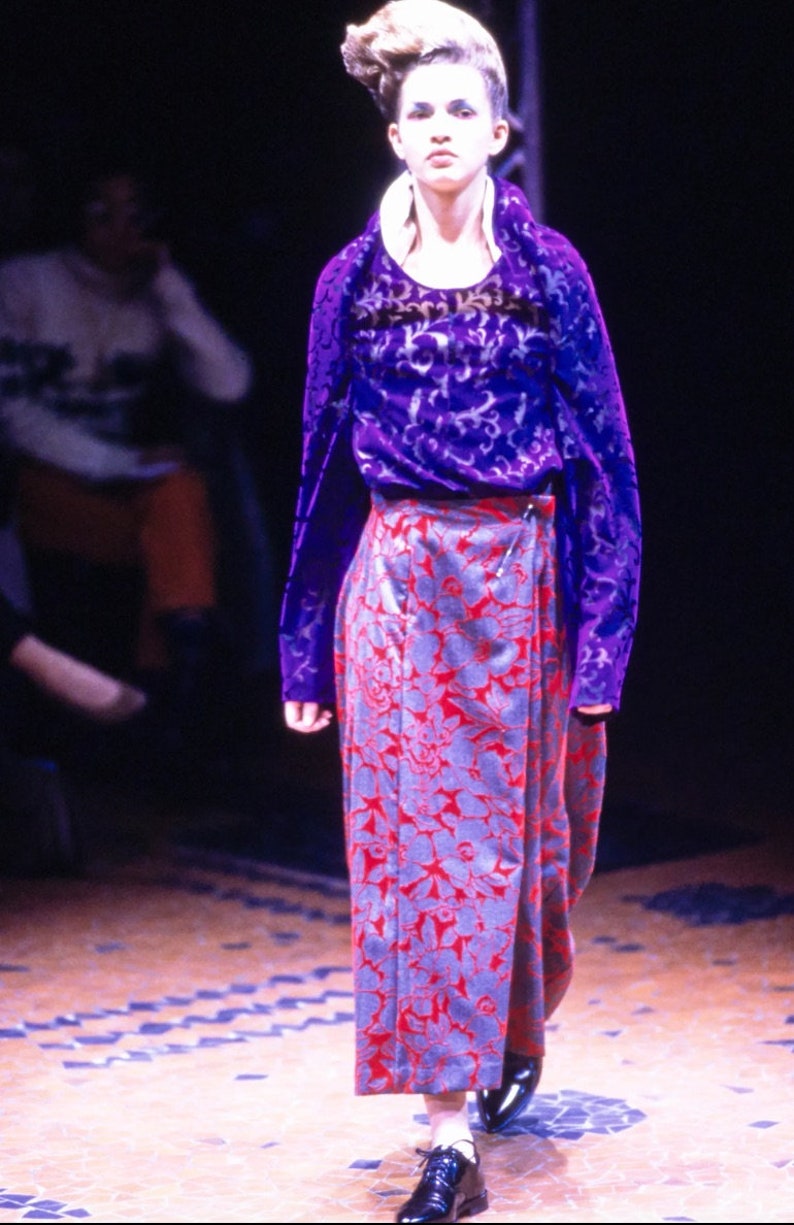 May include: A model wearing a purple and silver patterned top with a long, red and silver floral skirt. The skirt has a wrap-around style and is cinched at the waist with a belt.