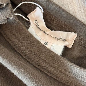 May include: A brown cotton t-shirt with a white tag that reads "Comme des Gar&ccedil;ons" and "S" in black text. The tag also has the text "Japan" in black text.