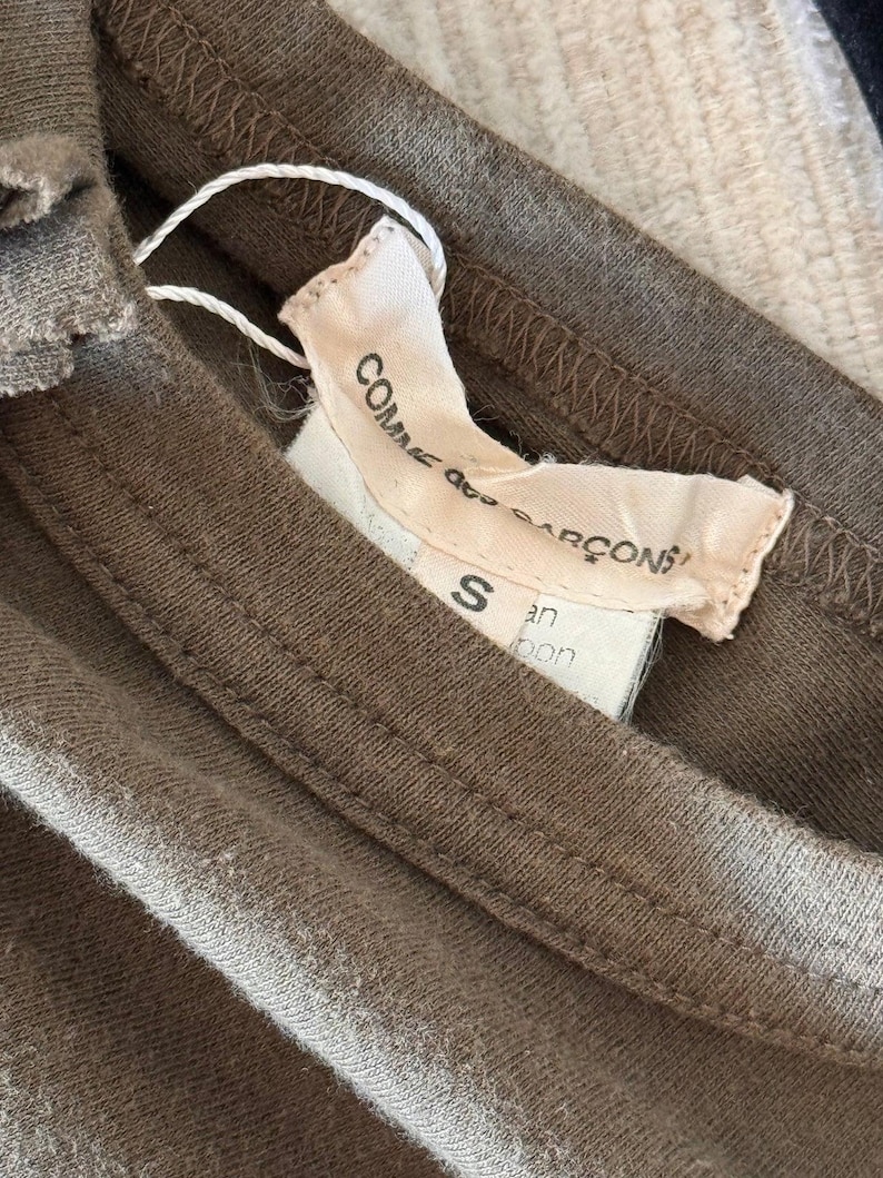 May include: A brown cotton t-shirt with a white tag that reads "Comme des Gar&ccedil;ons" and "S" in black text. The tag also has the text "Japan" in black text.