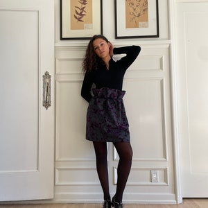 May include: A woman wearing a black mock neck shirt, a purple and gray floral skirt, black tights, and black shoes. She is standing in front of a white wall with two framed botanical prints.