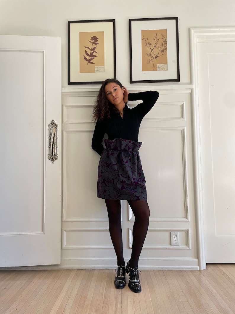 May include: A woman wearing a black mock neck shirt, a purple and gray floral skirt, black tights, and black shoes. She is standing in front of a white wall with two framed botanical prints.