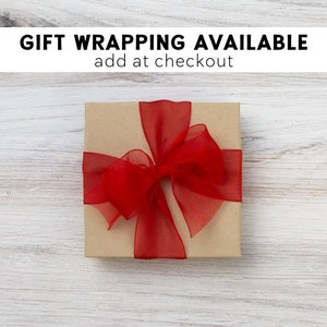 May include: A brown gift box with a red ribbon and bow. The box is on a white wooden surface. The text "GIFT WRAPPING AVAILABLE add at checkout" is above the box.
