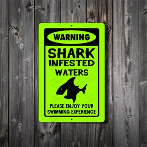 May include: A neon green rectangular sign with black text and a shark silhouette. The sign reads "WARNING SHARK INFESTED WATERS" above a shark graphic. Below the graphic, it says "PLEASE ENJOY YOUR SWIMMING EXPERIENCE".