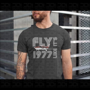 May include: A dark gray t-shirt with a white graphic design that says "FLY LIKE IT'S 1977" with a small airplane image in the middle of the text.