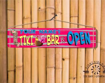 May include: A rectangular wooden sign with the text "YOUR NAME'S TIKI BAR IS OPEN" in blue, yellow, and pink. The sign features a tiki mask, a cocktail, and floral accents. It hangs against a bamboo background.