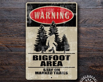 May include: A rectangular metal sign with a distressed finish. The sign features a red "WARNING" oval, a Bigfoot silhouette, and the text "BIGFOOT AREA STAY ON MARKED TRAILS". The sign is set against a wooden background.